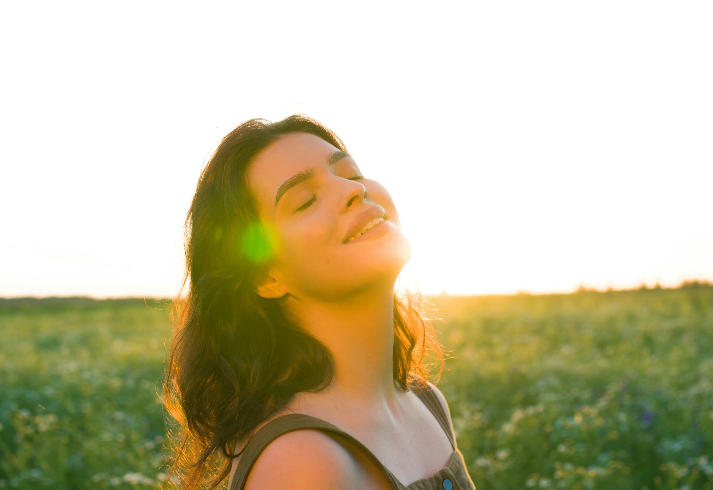 A woman with eyes closed, smiling in warm sunlight in a green meadow